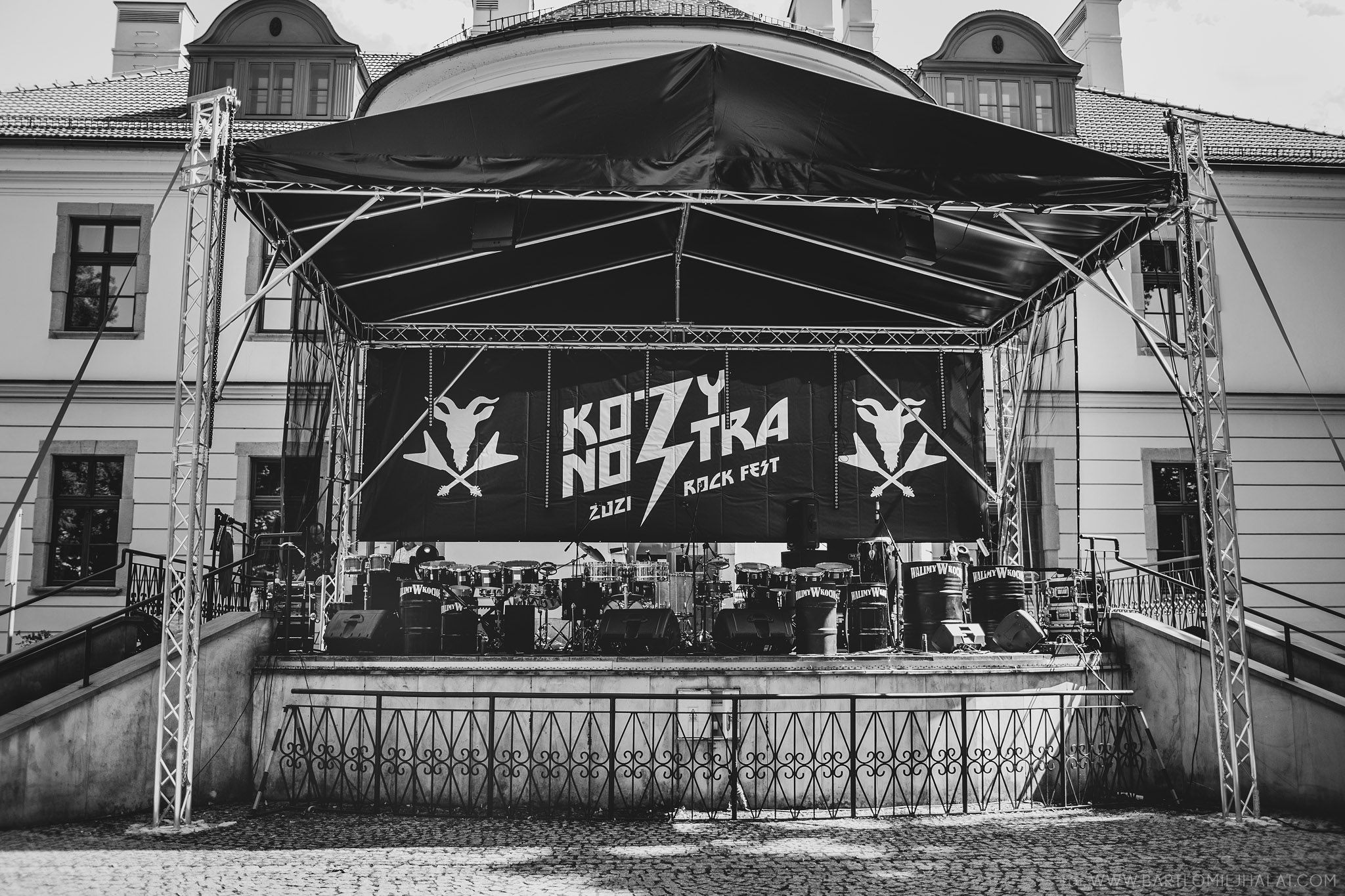 Czarno-biała fotografia przedstawiająca przygotowaną scenę KozyNostra Rock Fest 2021 przed rozpoczęciem koncertów. Na tarasie Pałacu Czeczów w Kozach ustawiona jest zadaszona scena z banerem festiwalowym, zestawem instrumentów perkusyjnych oraz nagłośnieniem. W tle widoczna jest zabytkowa fasada pałacu. Zdjęcie ukazuje miejsce wydarzenia tuż przed pojawieniem się artystów i publiczności.