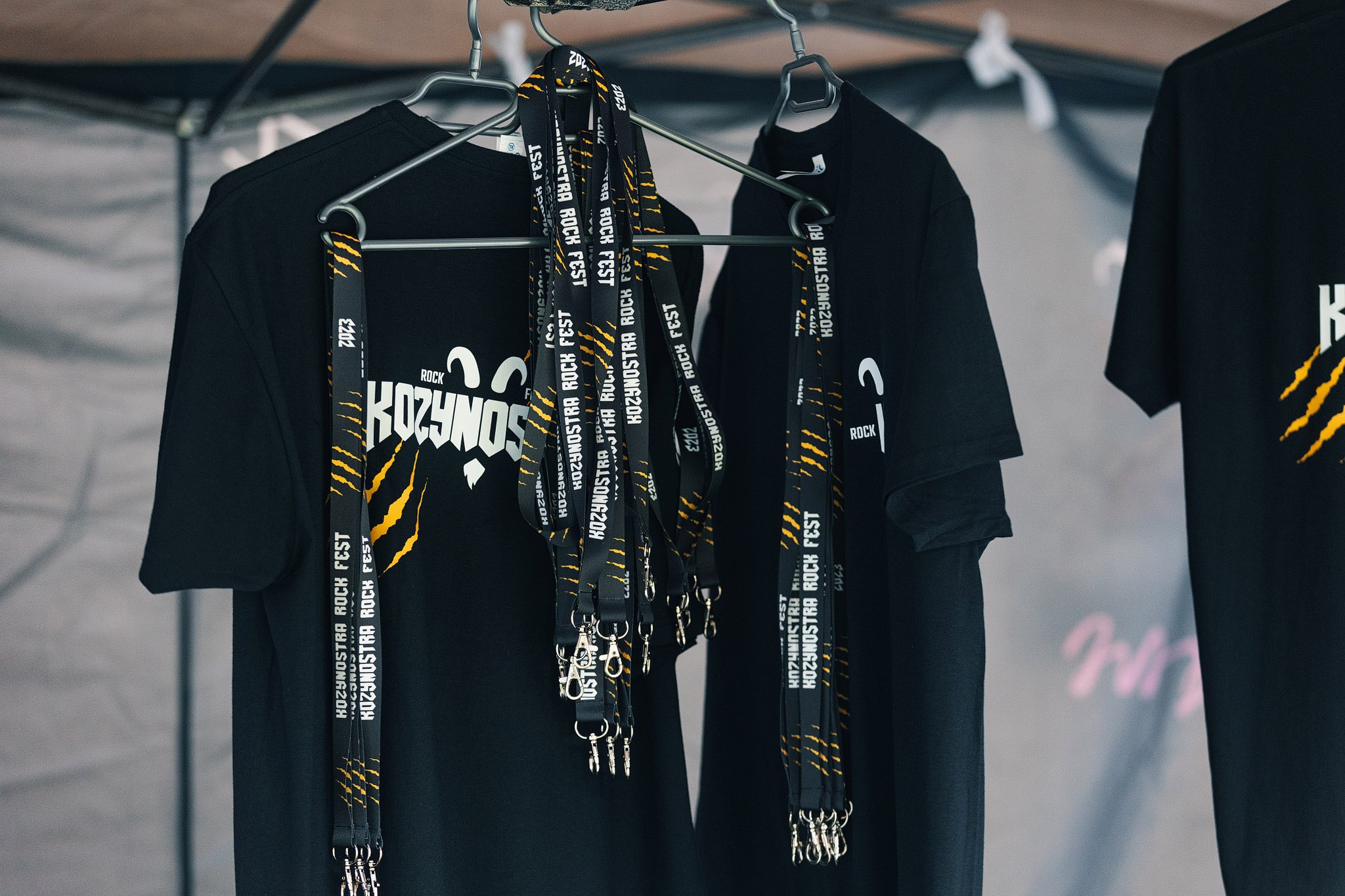 Na stoisku z festiwalowym merchem wiszą czarne koszulki z logo KozyNostra Rock Fest oraz smycze z nazwą festiwalu przeznaczone do sprzedaży uczestnikom wydarzenia.