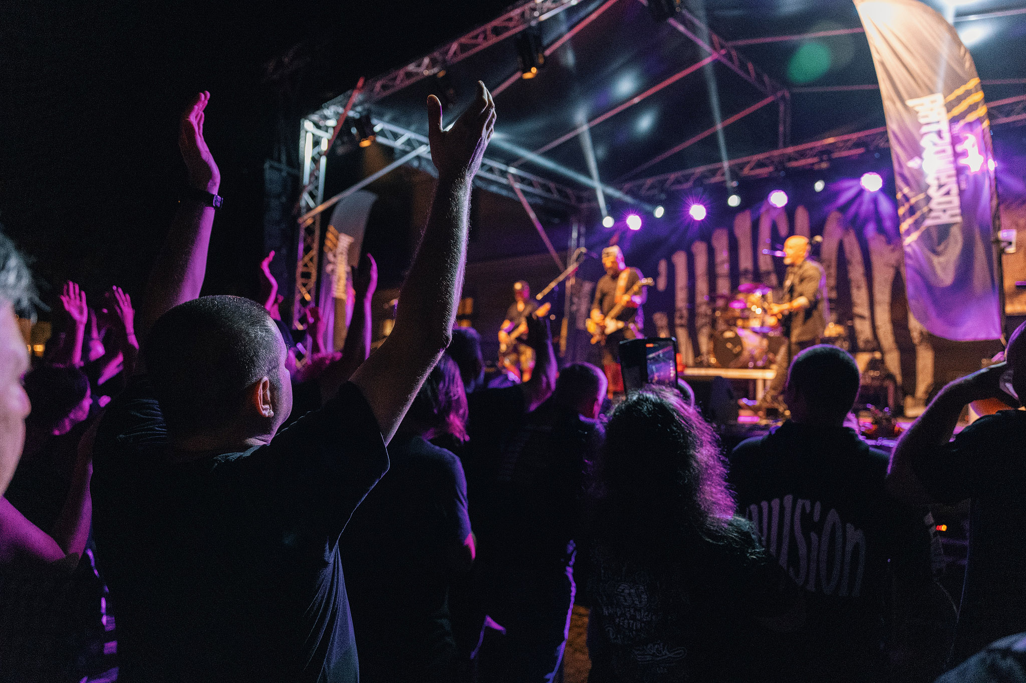 Tłum uczestników KozyNostra Rock Fest 2024 bawi się pod sceną podczas wieczornego koncertu. W górze uniesione ręce publiczności, kolorowe światła oświetlają scenę z występującym zespołem, a po bokach widoczne są flagi z logo festiwalu, oddające energię i atmosferę rockowego wydarzenia.