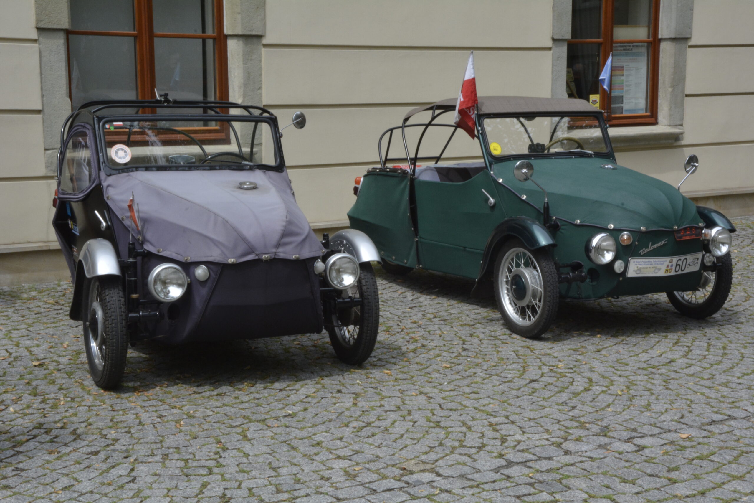 Na brukowanym dziedzińcu przed budynkiem stoją dwa zabytkowe trójkołowe pojazdy Velorex. Po lewej stronie znajduje się ciemnofioletowy model z materiałowym nadwoziem, a po prawej zielony Velorex z otwartą konstrukcją i biało-czerwoną flagą zamocowaną z tyłu. W tle widoczna jest elewacja budynku z dużymi oknami. Zdjęcie przedstawia historyczne pojazdy prezentowane podczas wydarzenia motoryzacyjnego.