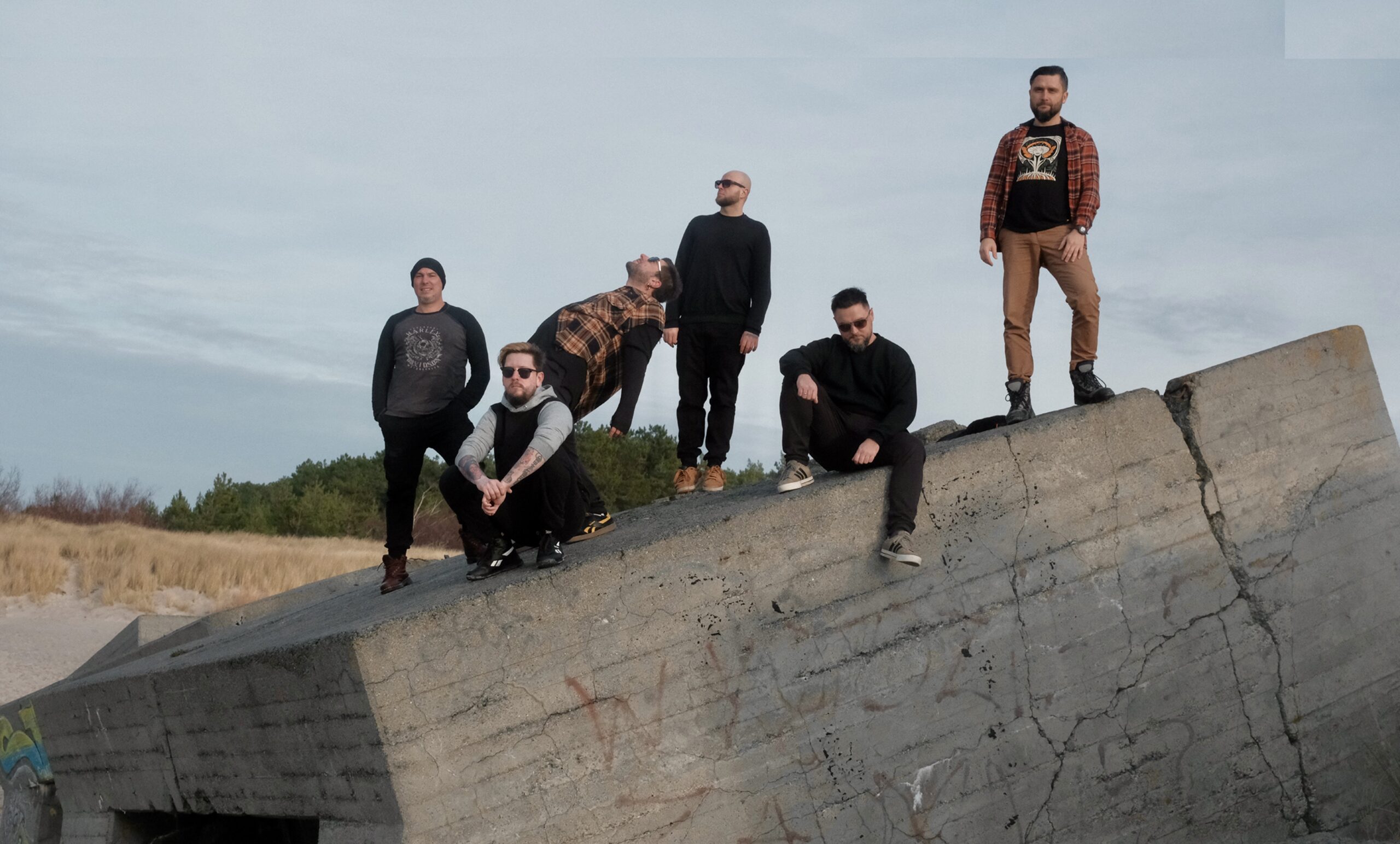 Sześciu członków zespołu rockowego pozuje na dużym betonowym bunkrze w nadmorskim krajobrazie. Muzycy stoją i siedzą w różnych pozach, ubrani w casualowe stroje w ciemnych kolorach. W tle widoczne są wydmy, drzewa oraz zachmurzone niebo. Fotografia wykonana na świeżym powietrzu podkreśla swobodny, rockowy charakter zespołu.