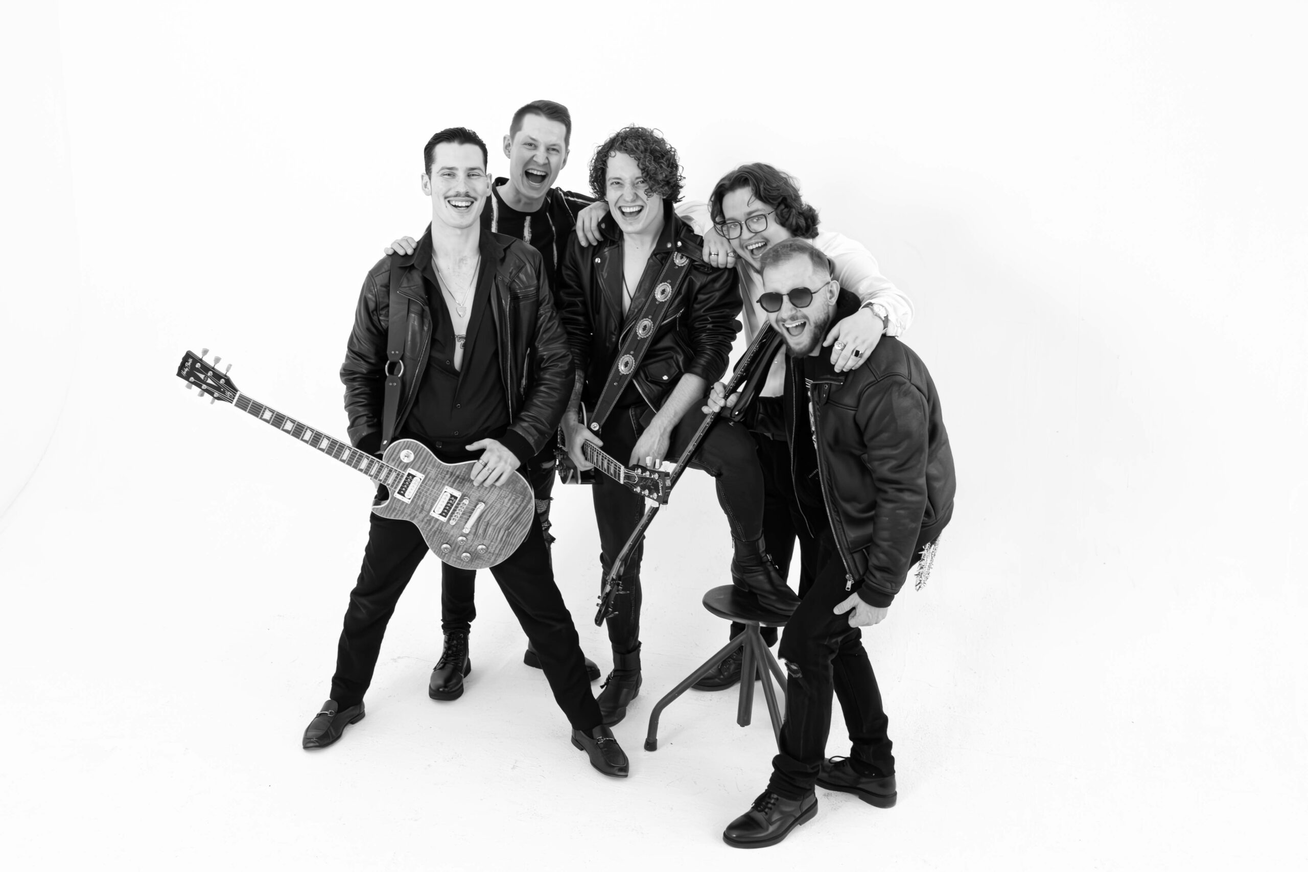 Pięciu członków zespołu rockowego pozuje do zdjęcia promocyjnego na jednolitym białym tle. Dwóch muzyków trzyma gitary elektryczne, pozostali stoją blisko siebie, uśmiechając się i obejmując ramionami. Ubrani są głównie w czarne stroje i skórzane kurtki. Czarno-biała fotografia oddaje energię, radość i sceniczny charakter zespołu.