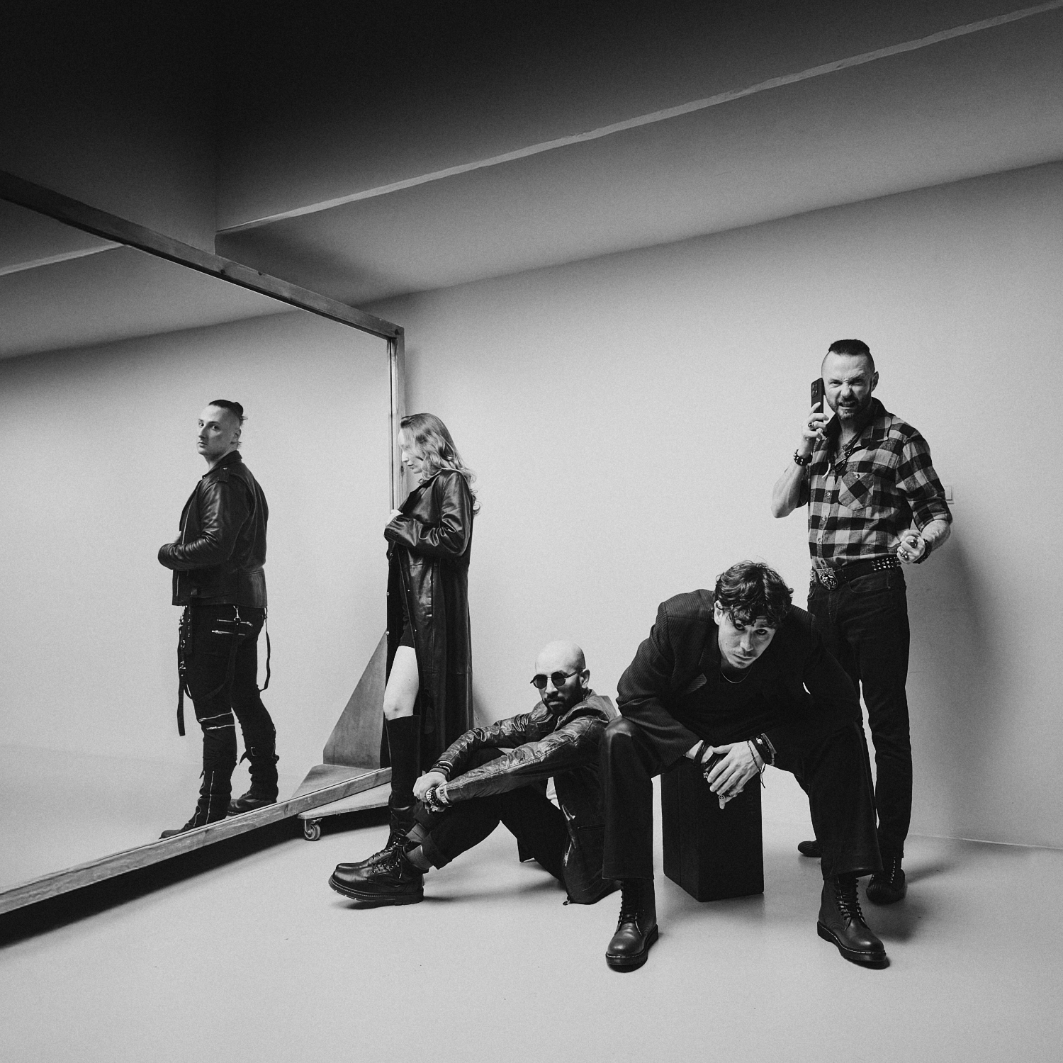 Czarno-białe zdjęcie promocyjne zespołu rockowego. Pięcioro muzyków pozuje w minimalistycznym studiu. Dwie osoby siedzą z przodu, dwie stoją z tyłu, a jedna widoczna jest przy dużym lustrze ustawionym z lewej strony kadru. Członkowie zespołu ubrani są głównie na czarno, w skórzane kurtki i ciężkie buty. Fotografia utrzymana jest w nowoczesnej, kontrastowej estetyce, podkreślającej rockowy charakter zespołu.