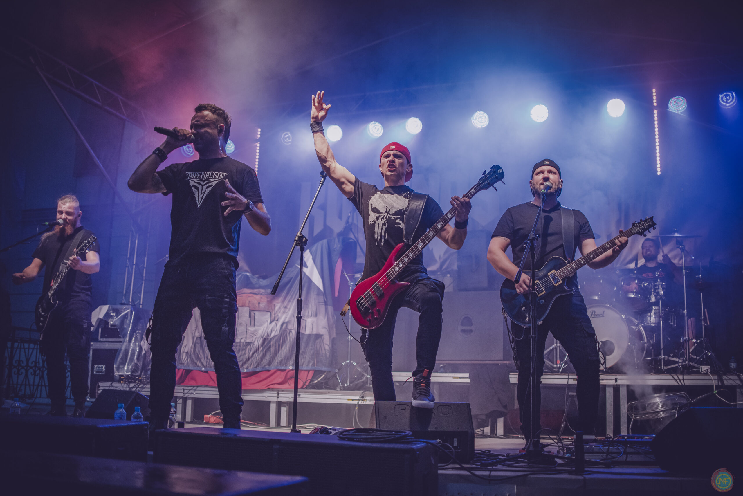 Pięcioosobowy zespół rockowo-metalowy występuje na scenie podczas KozyNostra Rock Fest 2022. Wokalista śpiewa do mikrofonu, obok gitarzyści i basista grają z dużą energią, a perkusista widoczny jest z tyłu sceny. Muzycy są oświetleni niebiesko-fioletowymi reflektorami i otoczeni scenicznym dymem, co podkreśla koncertową atmosferę wydarzenia.